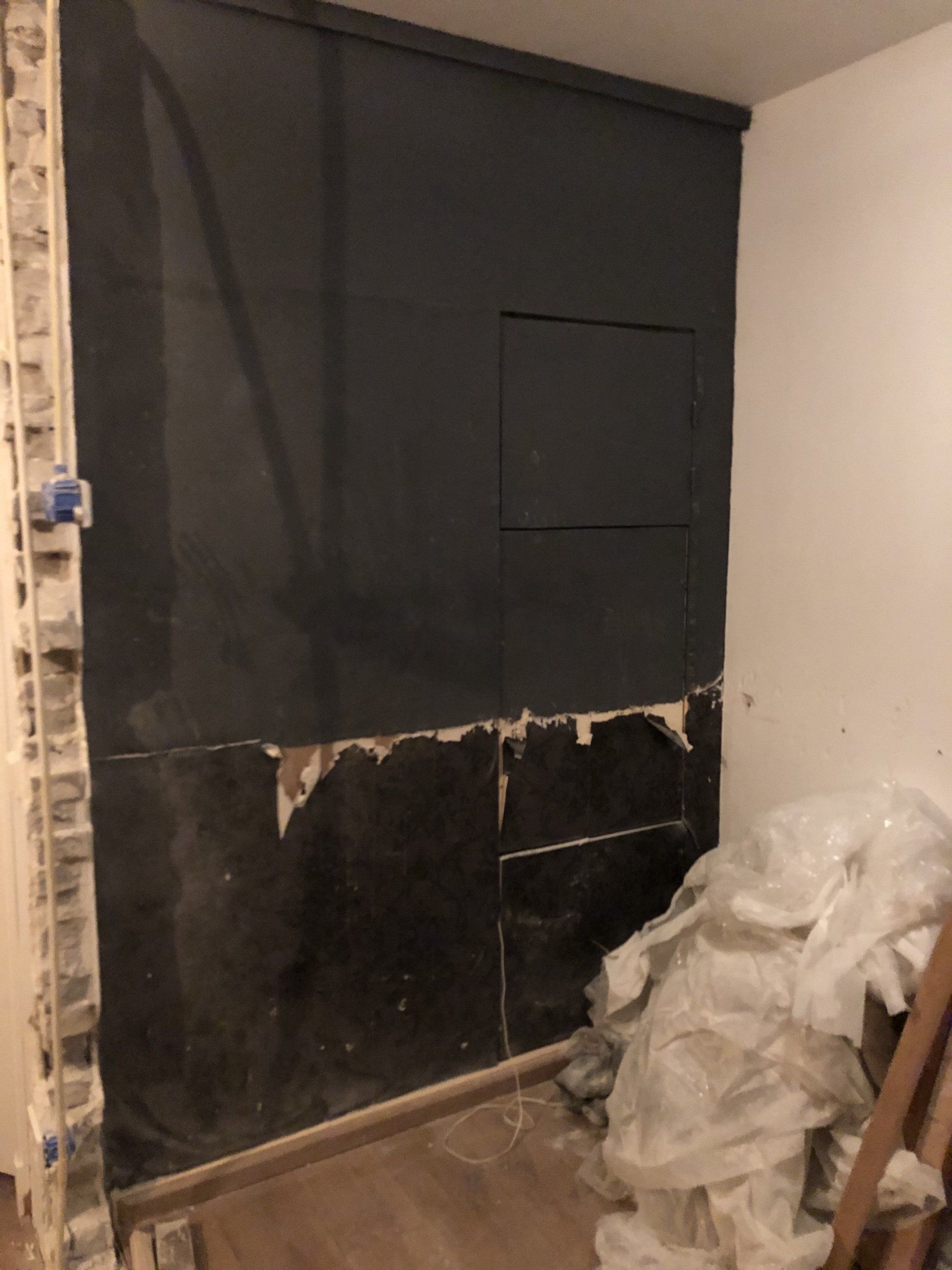 Muur met donker materiaal en een klein inspectieluik, in renovatie. Op de vloer liggen hopen puin.
