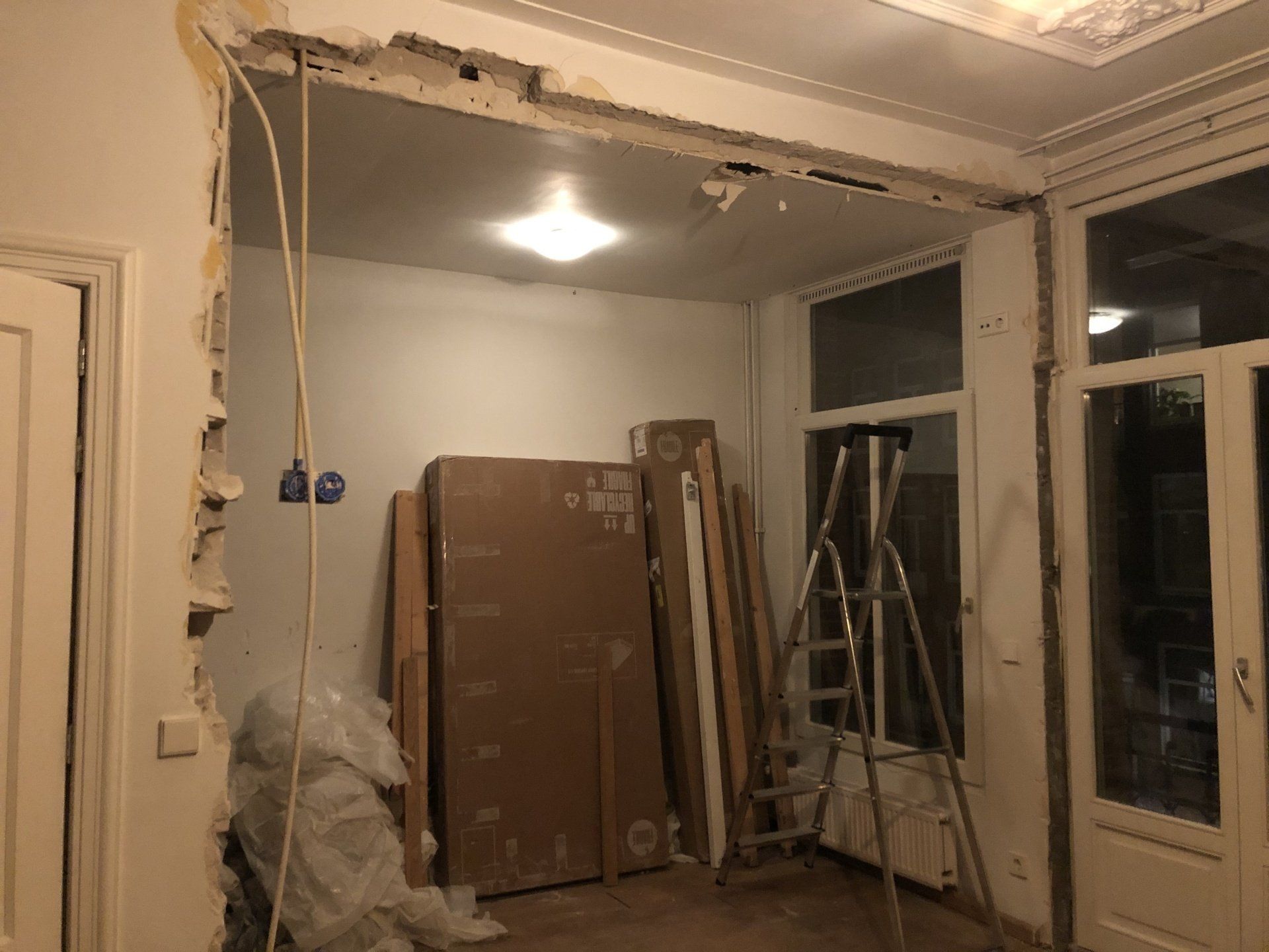 Renovatie in uitvoering: muur gedeeltelijk verwijderd, waardoor bedrading, plafondlamp en bouwmaterialen zichtbaar zijn.