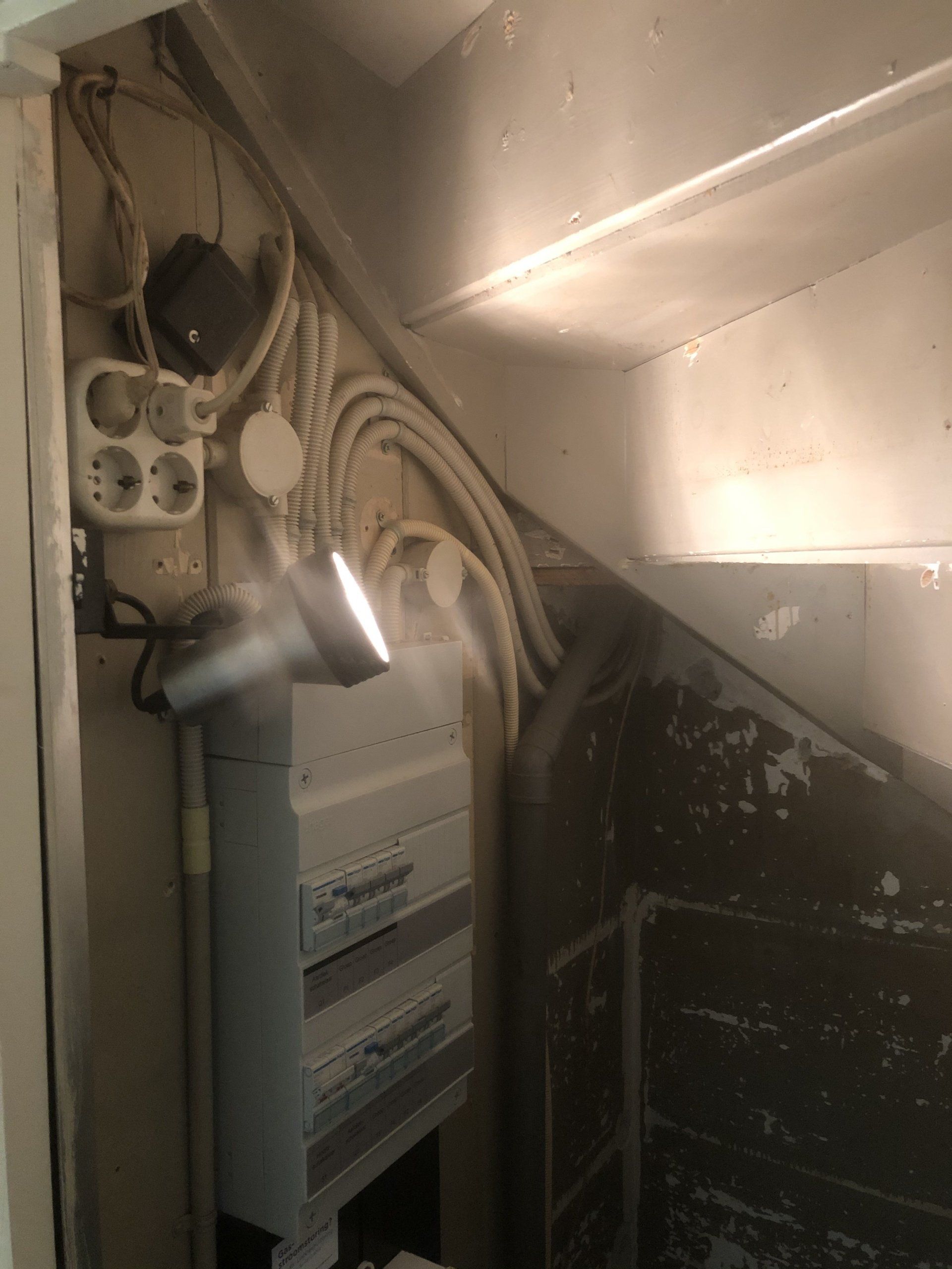 Elektrisch paneel en bedrading onder een trap. Inclusief stopcontacten, kabels en een spot. Grijze en witte tinten.