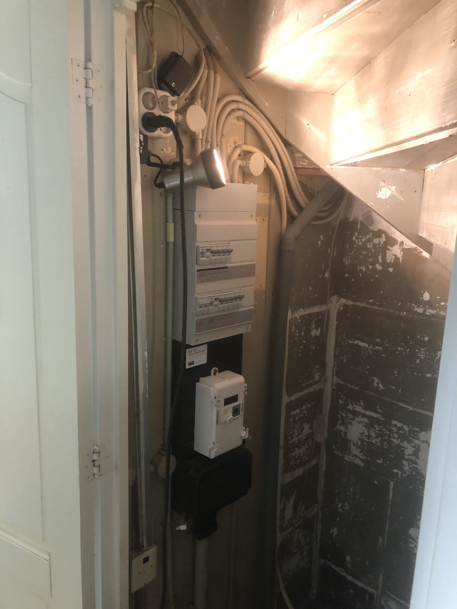 Elektrische meters en bedrading in een trappenhuis met zichtbaar beton en een wit deurkozijn.