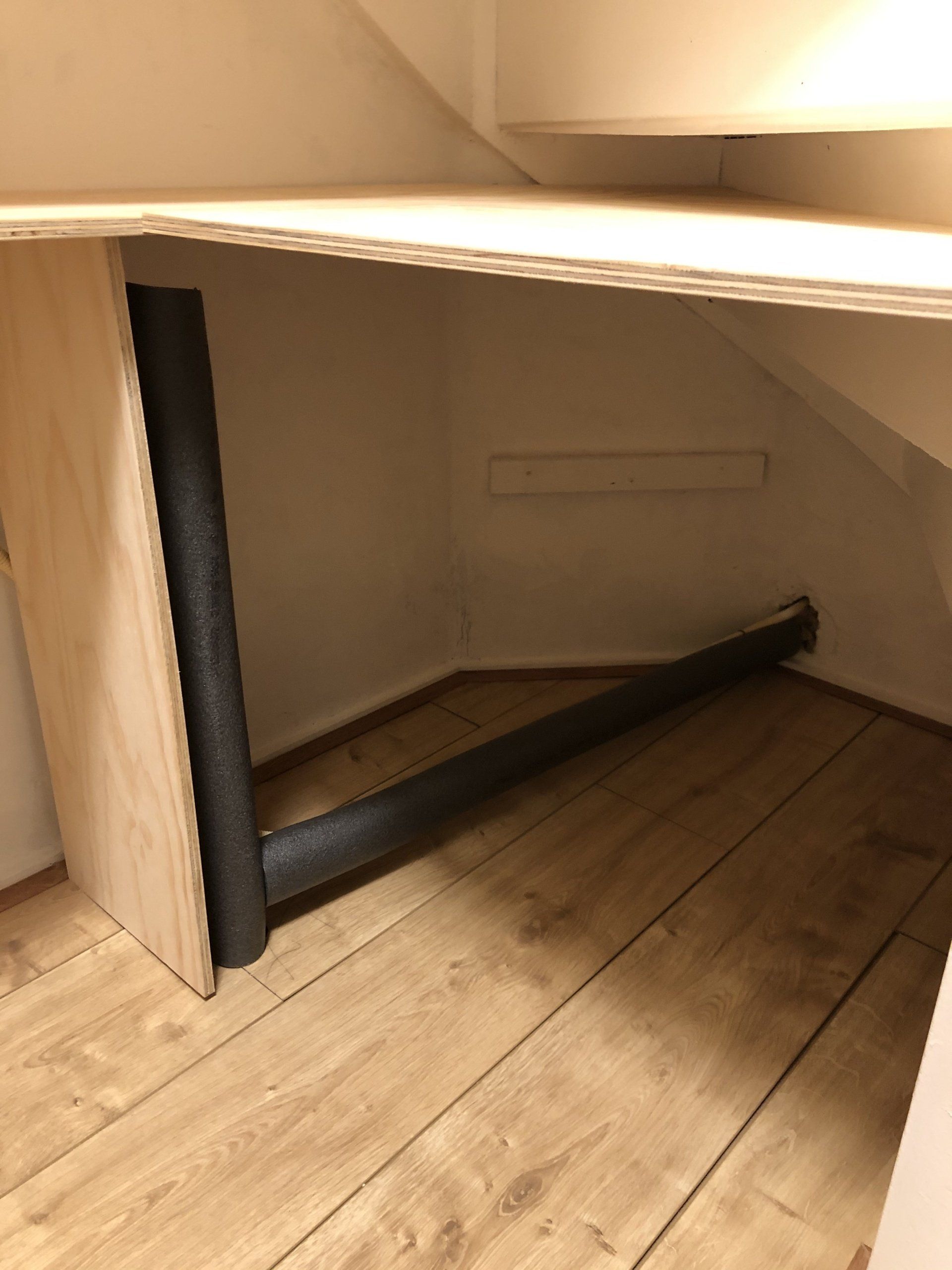 Houten opbergruimte onder de trap, met zichtbare leidingen en zwarte isolatie.