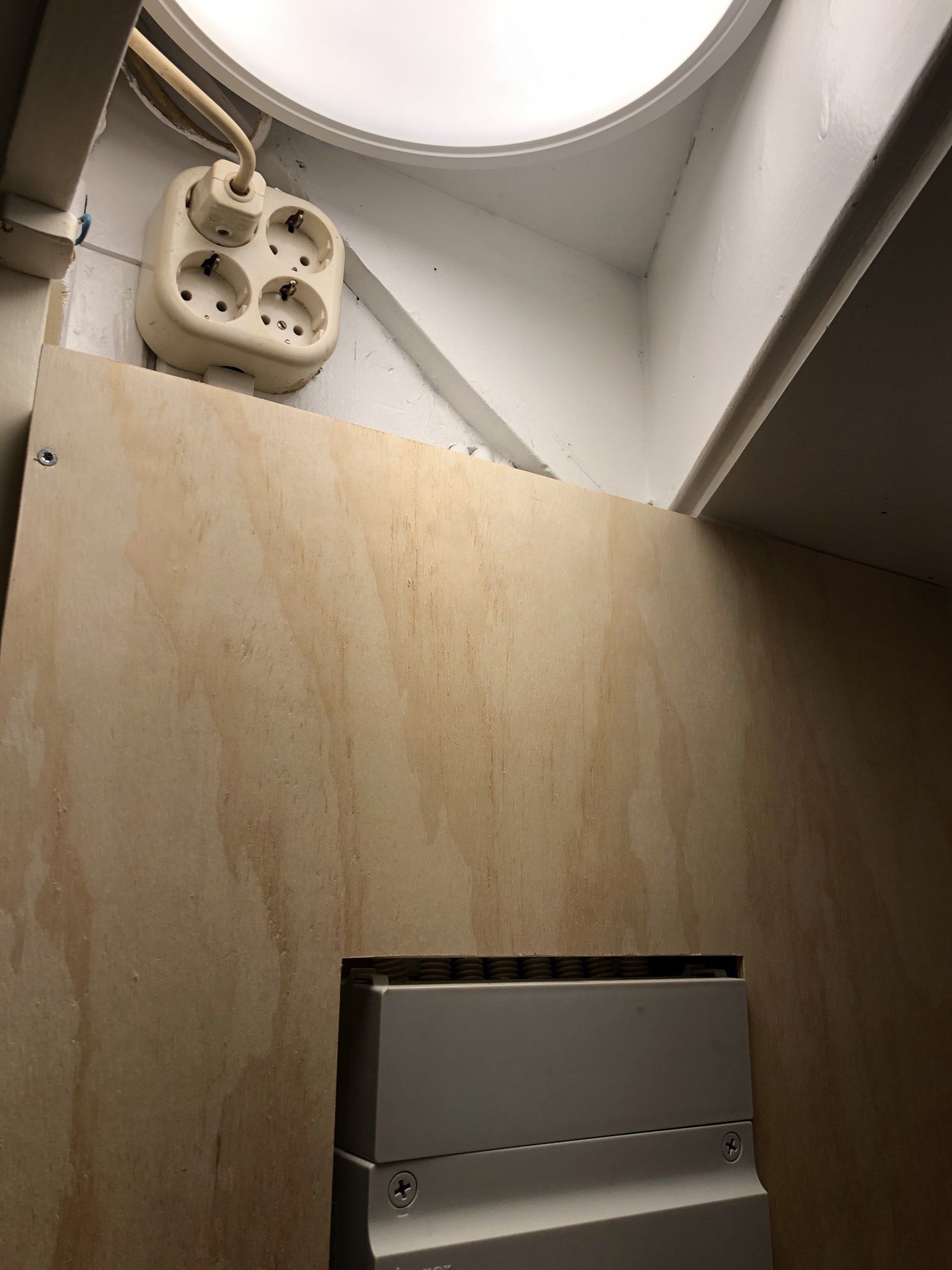 Houten muur met stopcontact, lamp zichtbaar in de bovenhoek. Er is een paneel of opening in de muur.