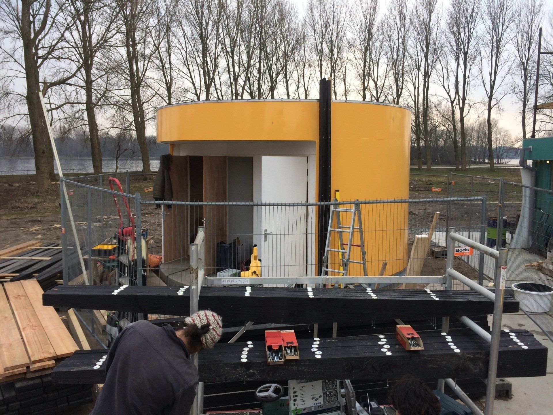 Persoon aan het werk aan een constructie; geel-wit gebouw op de achtergrond; bouwmaterialen op de voorgrond.
