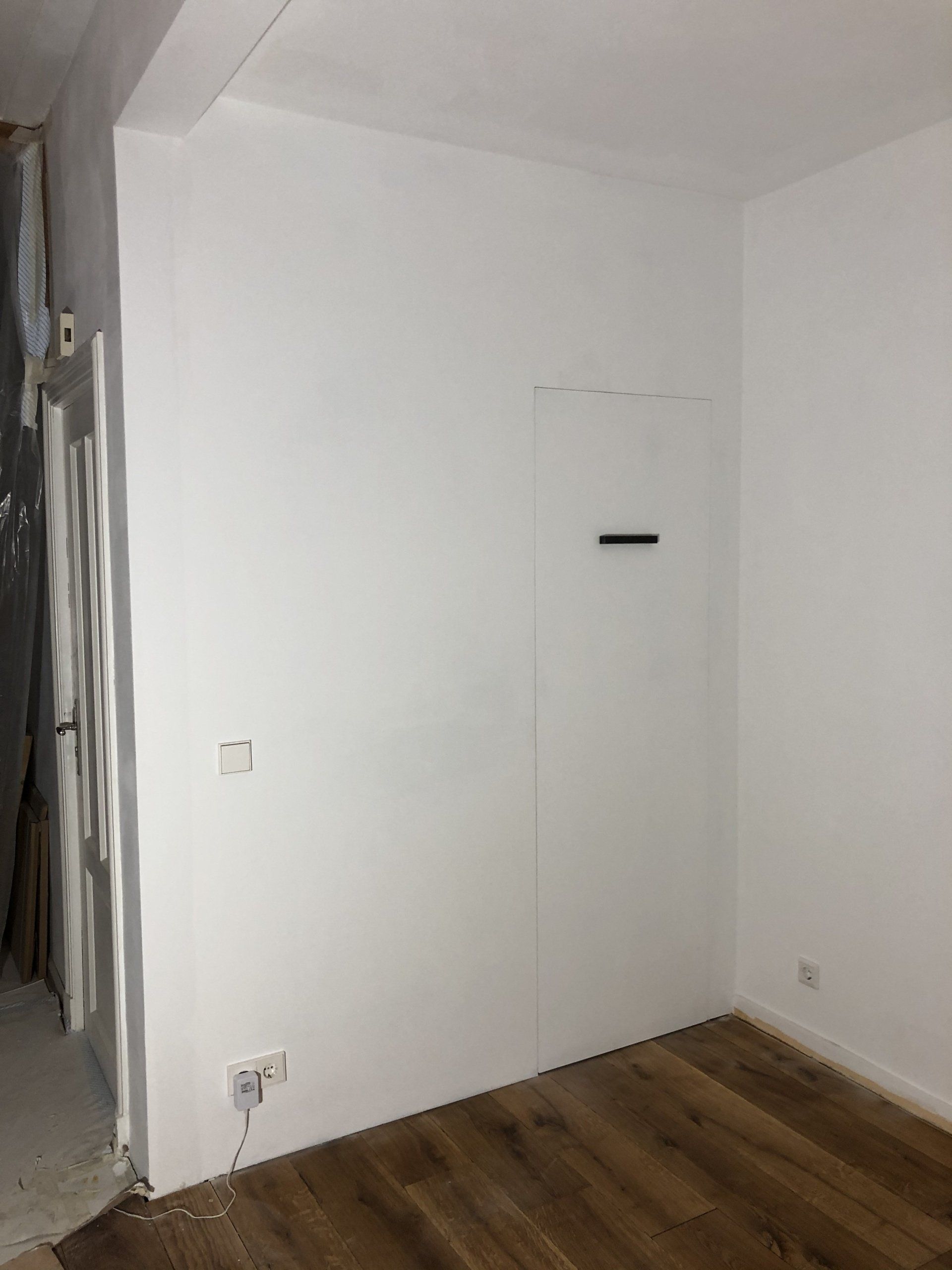 Witte muur met een deur, stopcontact en houten vloer. De kamer lijkt leeg.