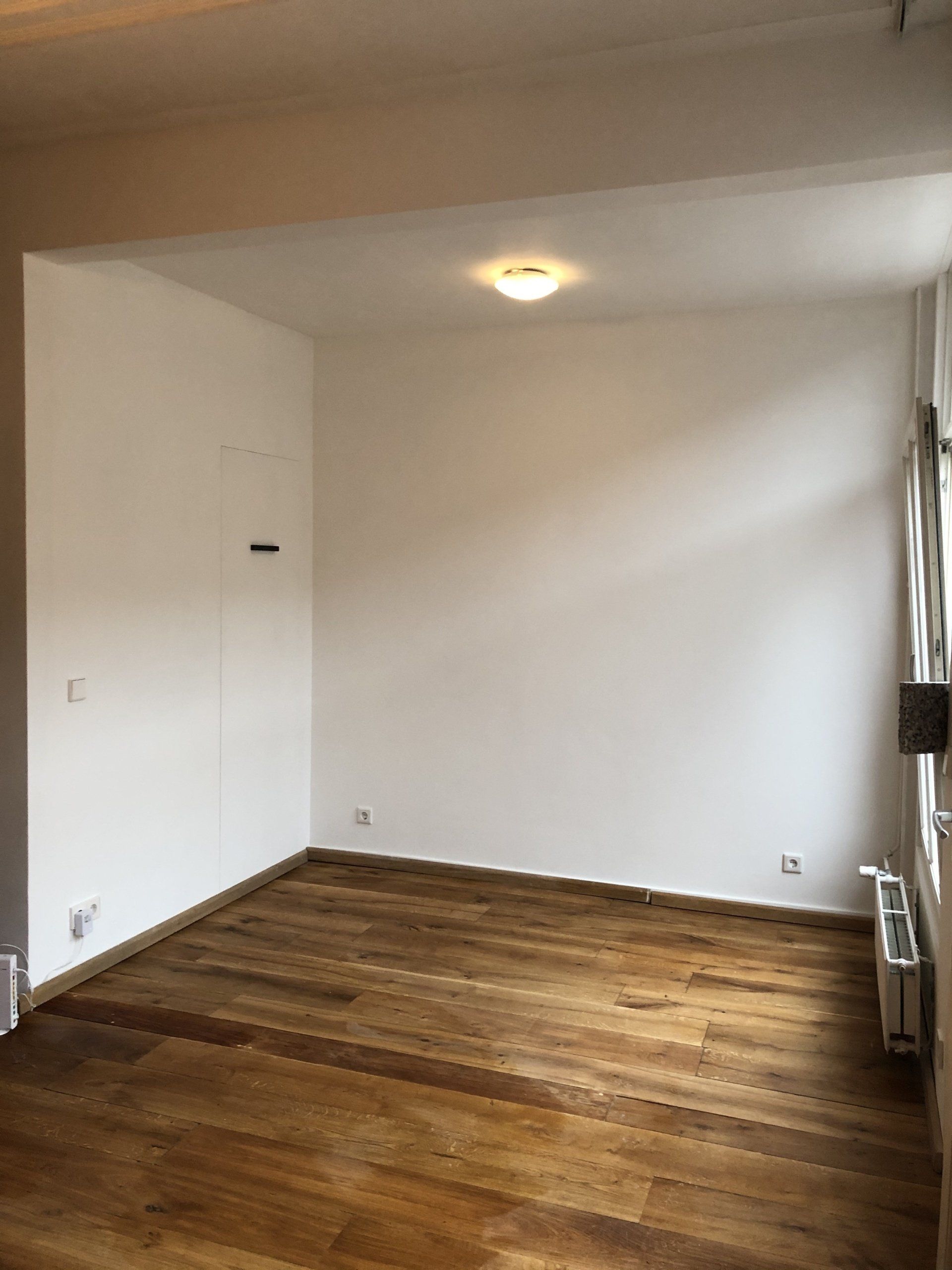 Een lege kamer met houten vloer, witte muren en een inbouwspot in het plafond.
