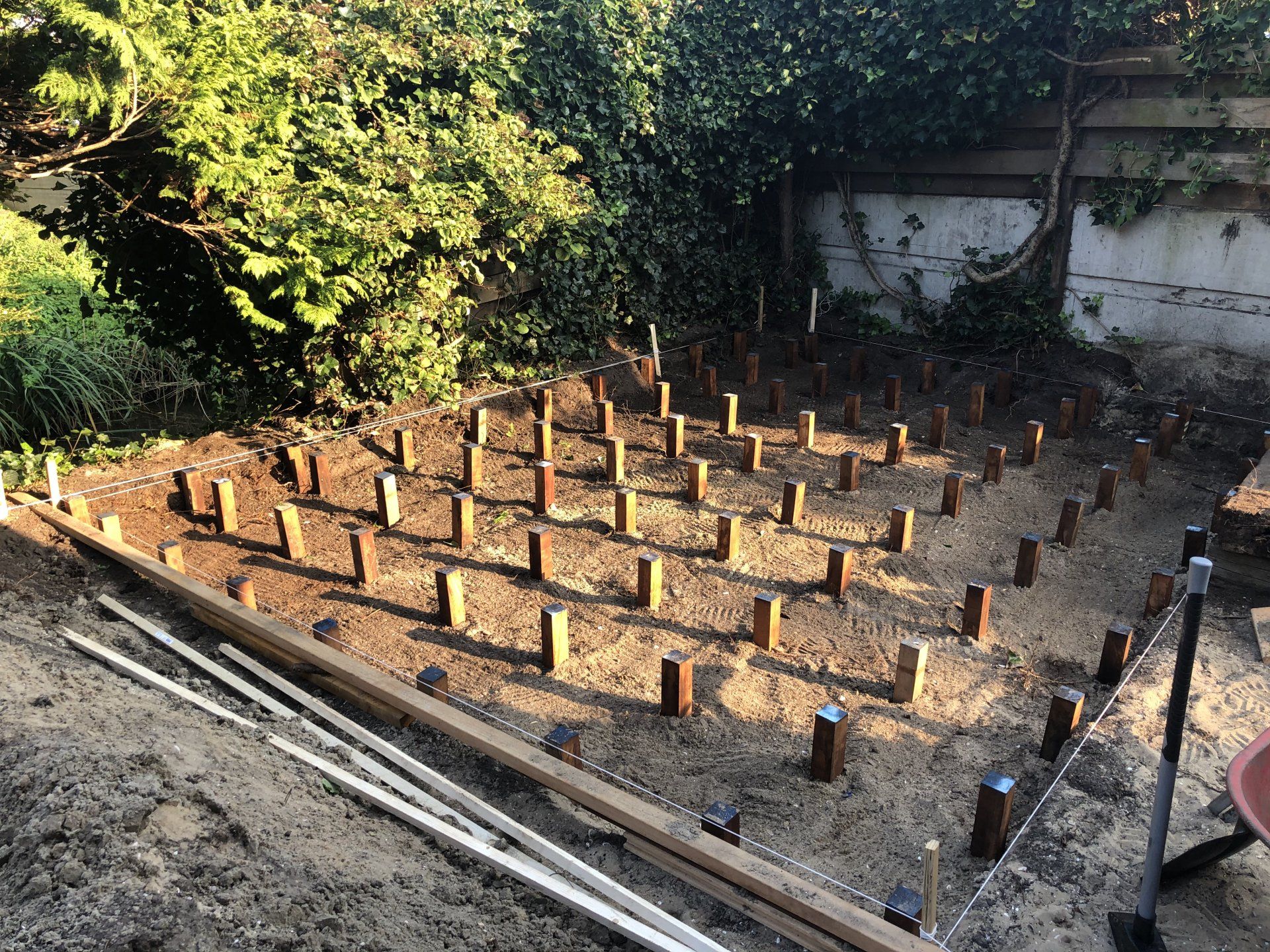 Houten palen in een voorbereid zandgebied, waarschijnlijk voor de aanleg van een terras. Omgeven door groen.