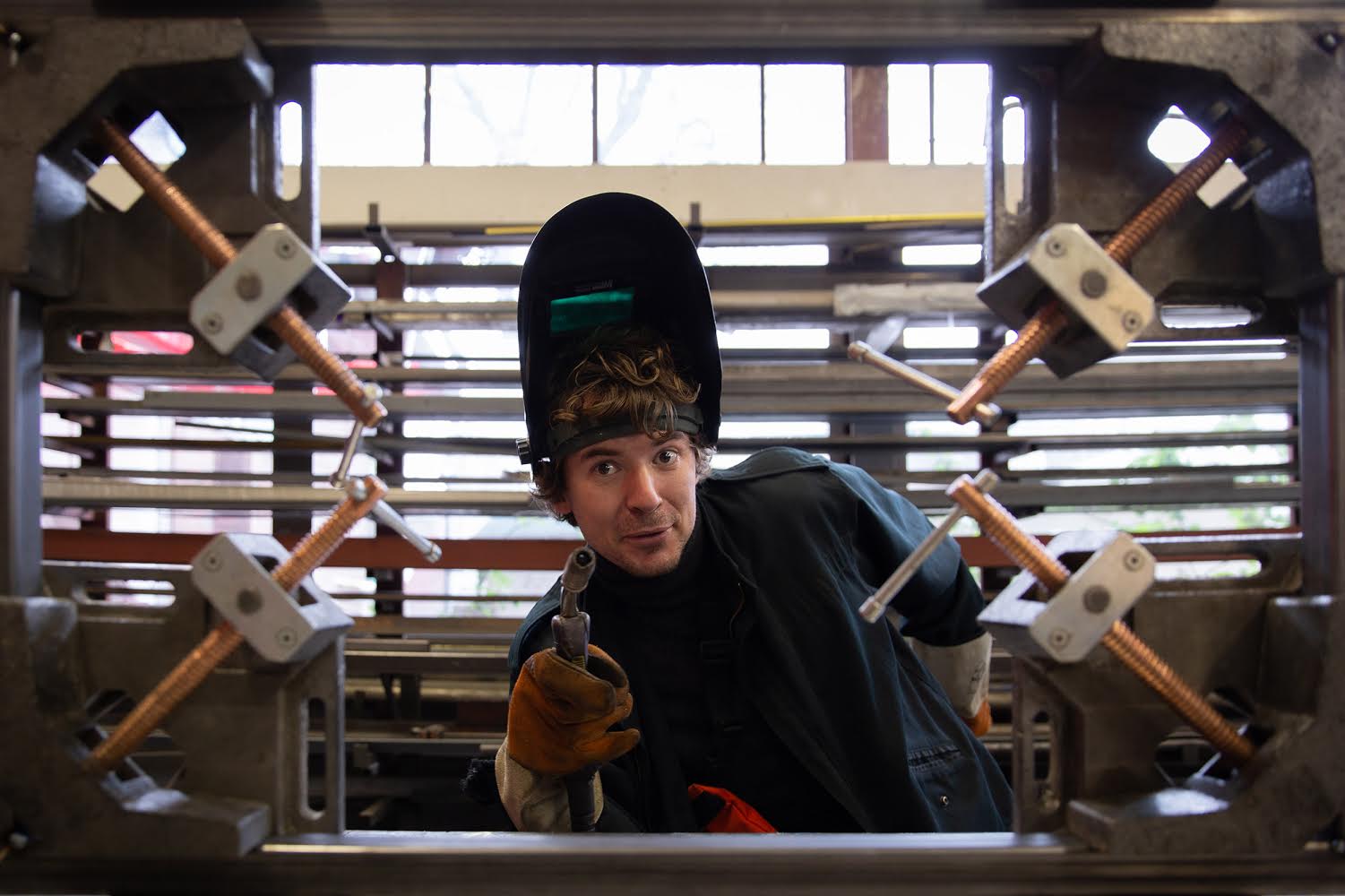 Lasser in beschermende kleding, wijst naar voren en lacht. Binnen een metalen frame met koperen staven, industriële omgeving.