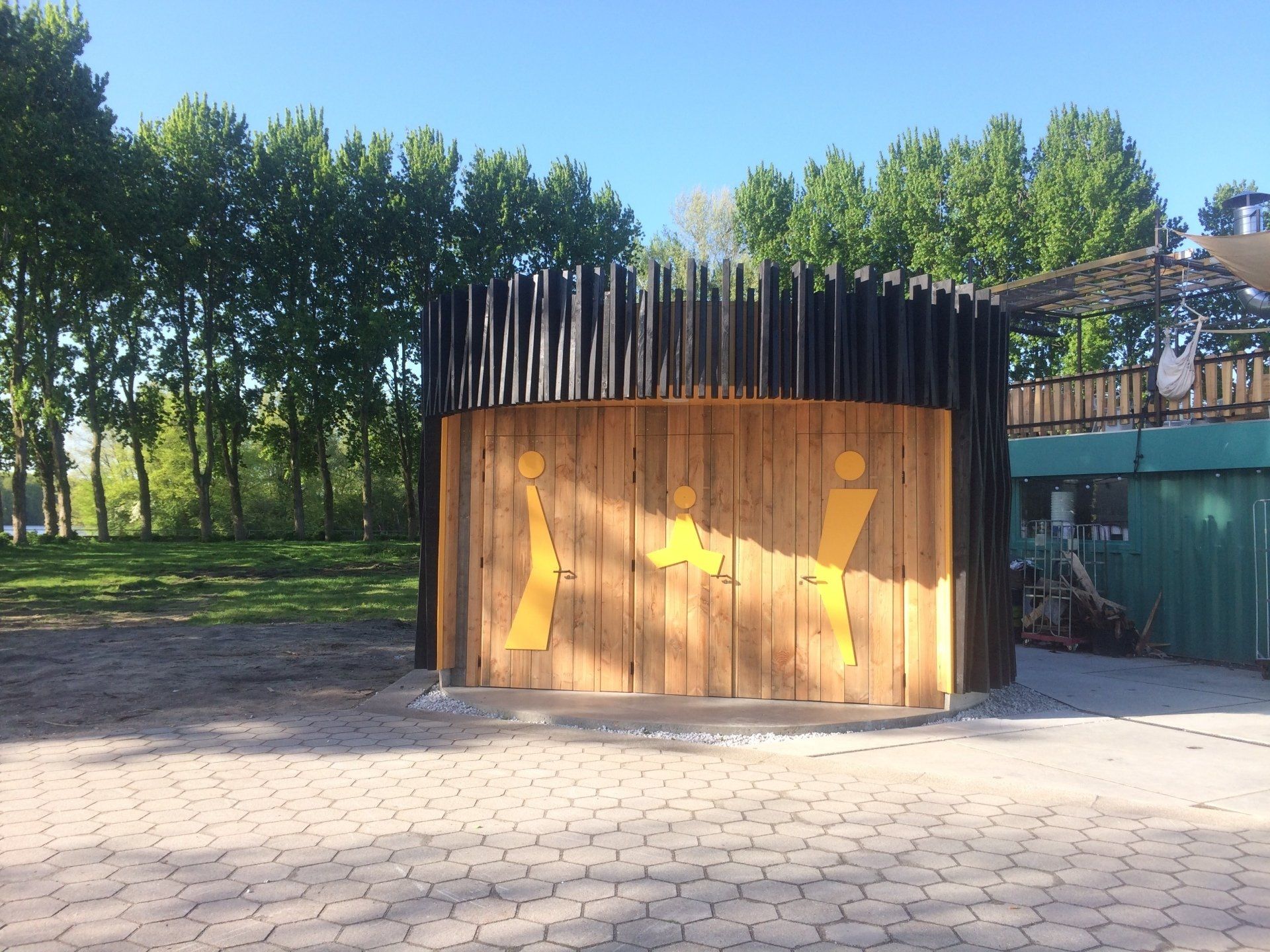 Houten constructie met gele uitgesneden figuren, omringd door donkere verticale palen, geplaatst op bestrating nabij bomen.
