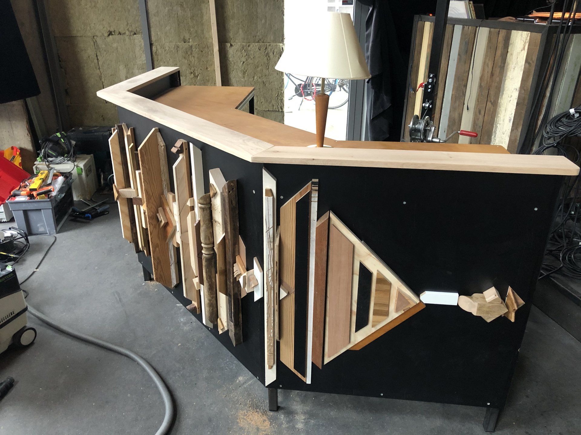 Een zwarte bar met geometrische houten versieringen, een lichthouten toonbank en een tafellamp in een rommelige werkplaats.