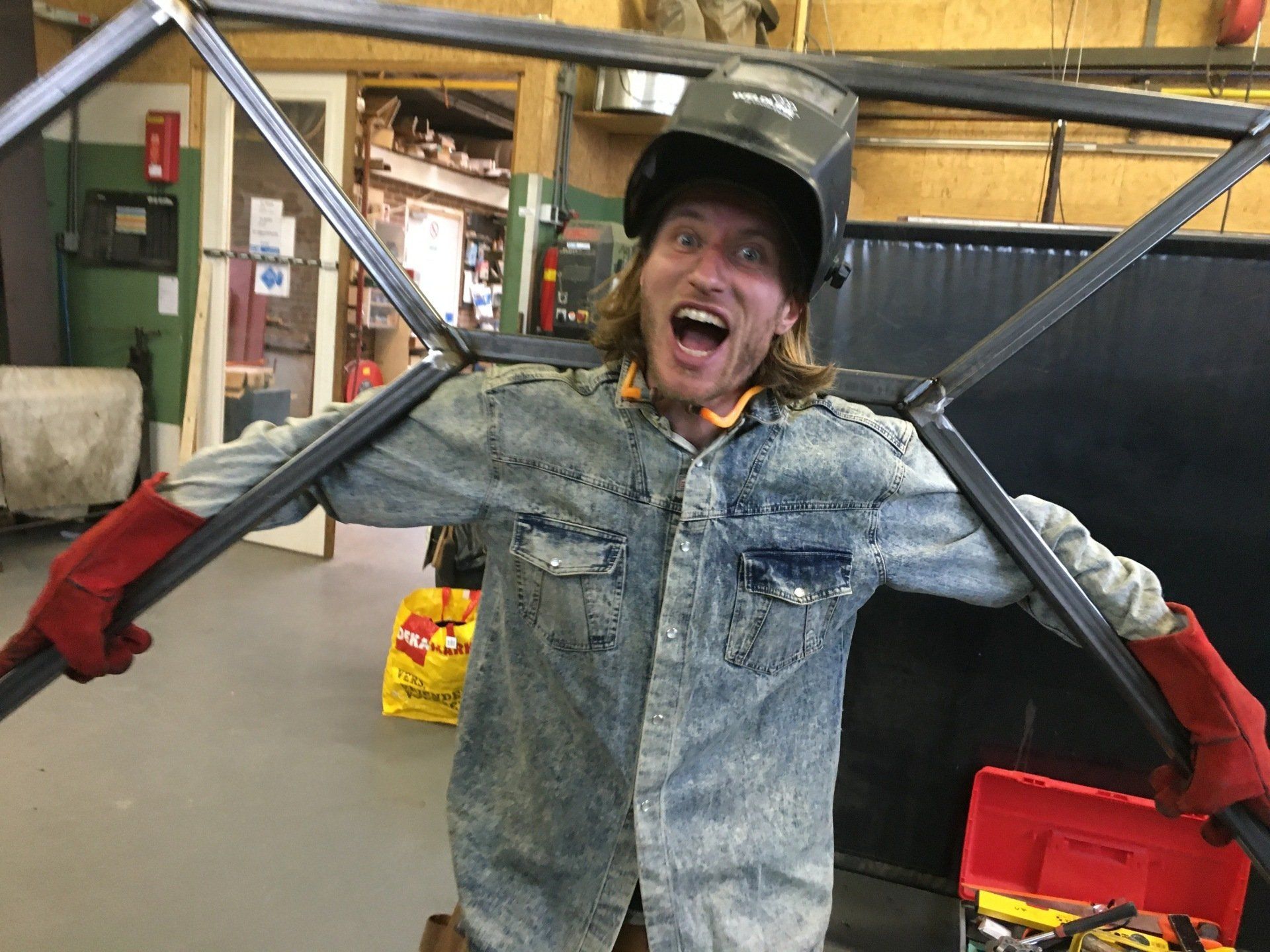 Een lasser in een spijkerjasje lacht, terwijl hij met rode lashandschoenen een metalen frame vasthoudt. Werkplaatsomgeving.