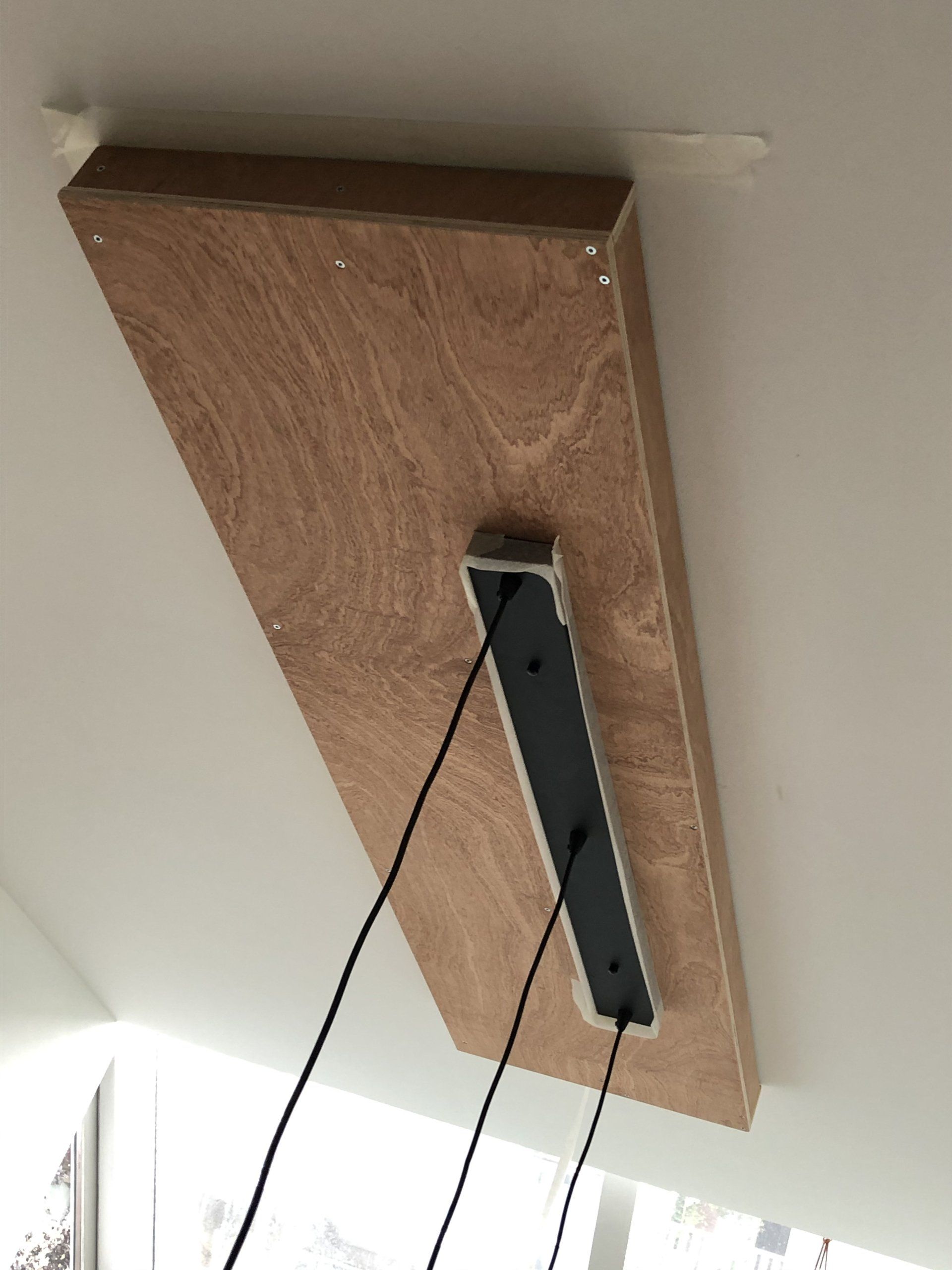 Een houten plank is aan het plafond bevestigd, waarop een rechthoekig armatuur met zwarte draden is gemonteerd.