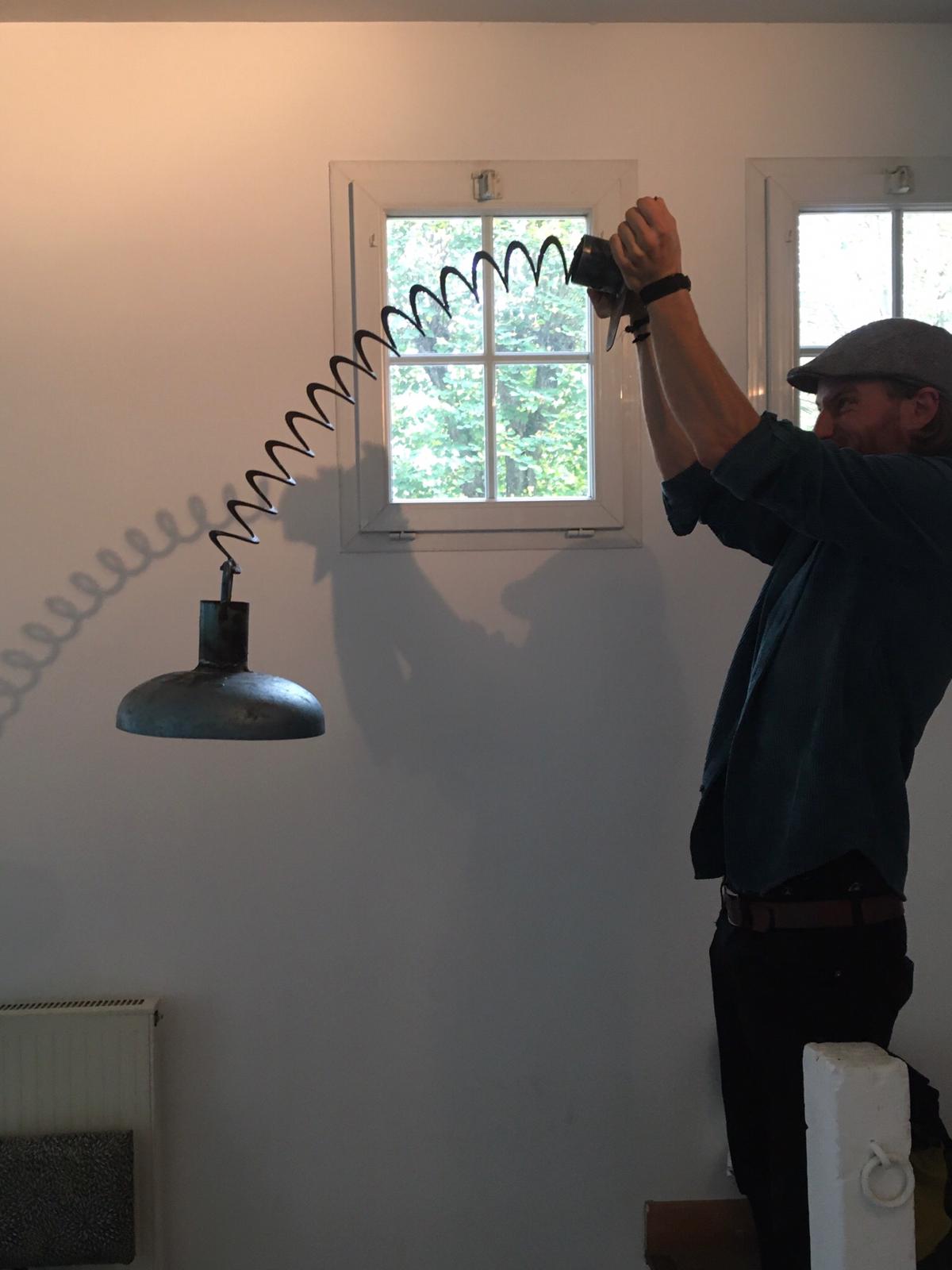 Een man installeert een hanglamp. De lamp hangt aan een opgerolde kabel vlakbij een raam; de man houdt de kabel omhoog.