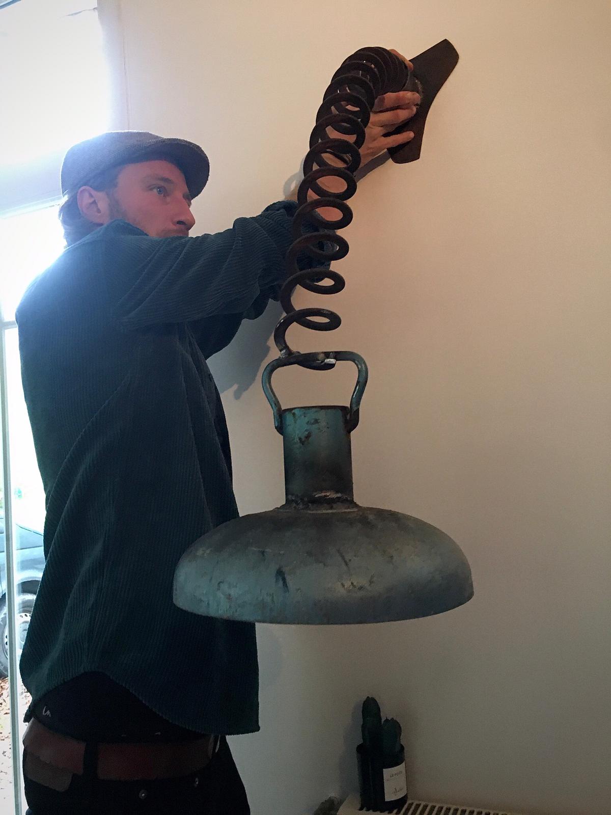 Een man installeert een grote, industriële lamp met een flexibele, opgerolde hals aan een witte muur.