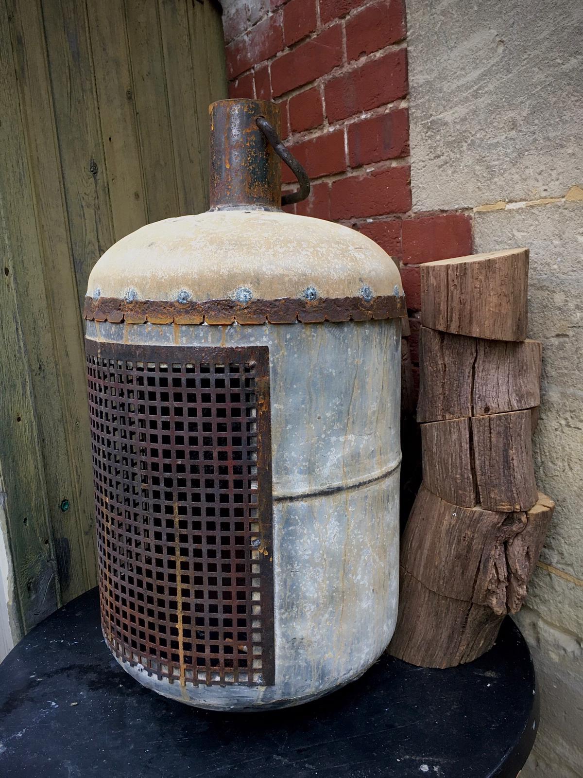 Metalen vat met draadgaas, naast brandhout, bakstenen muur.