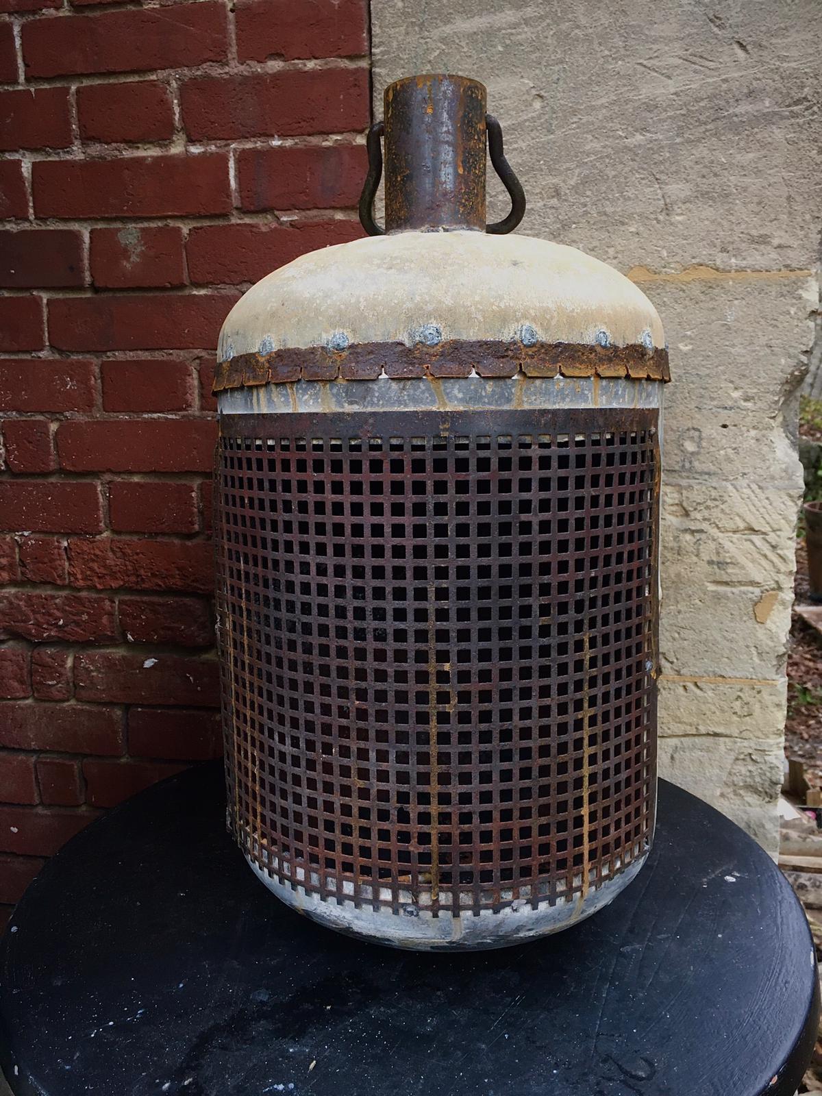 Een roestige metalen kooi met een betonnen bovenkant en handvat, die op een zwarte ondergrond naast een bakstenen muur staat.