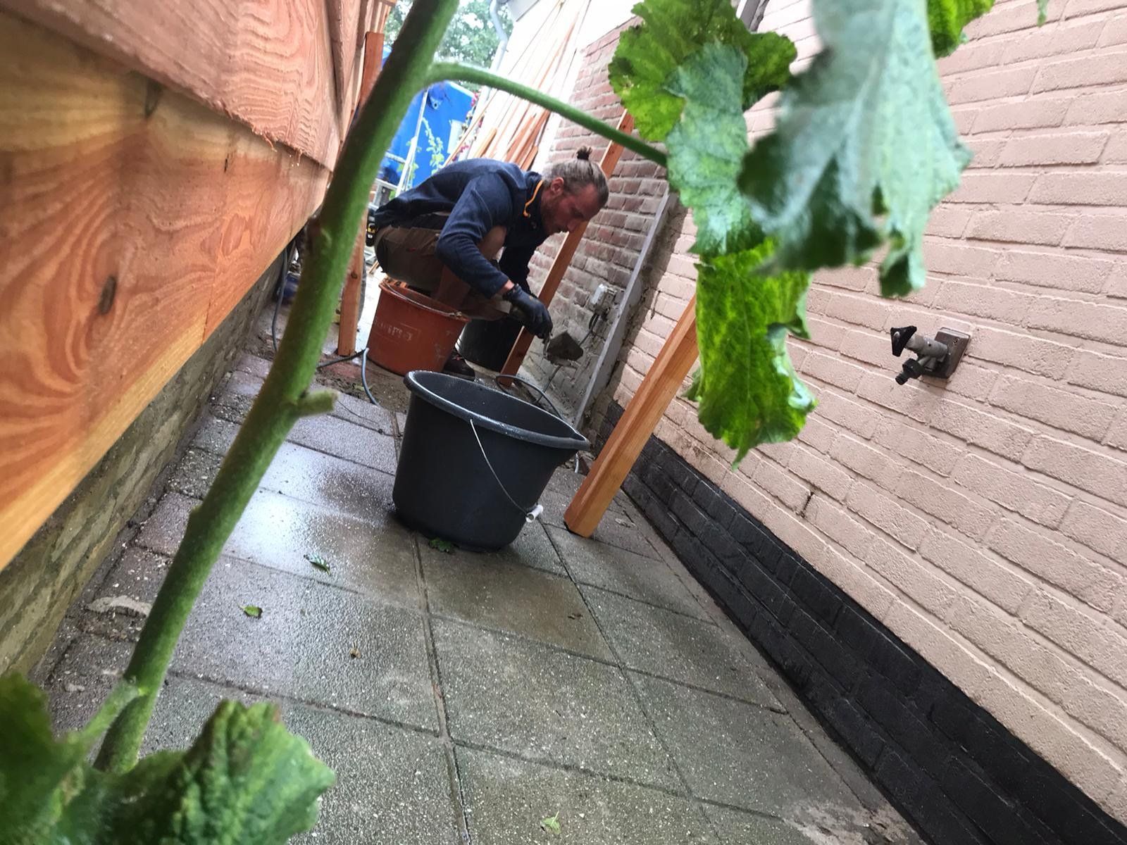 Een man in een blauw shirt staat in een smalle steeg bij een emmer, met een pompoenplant op de voorgrond.