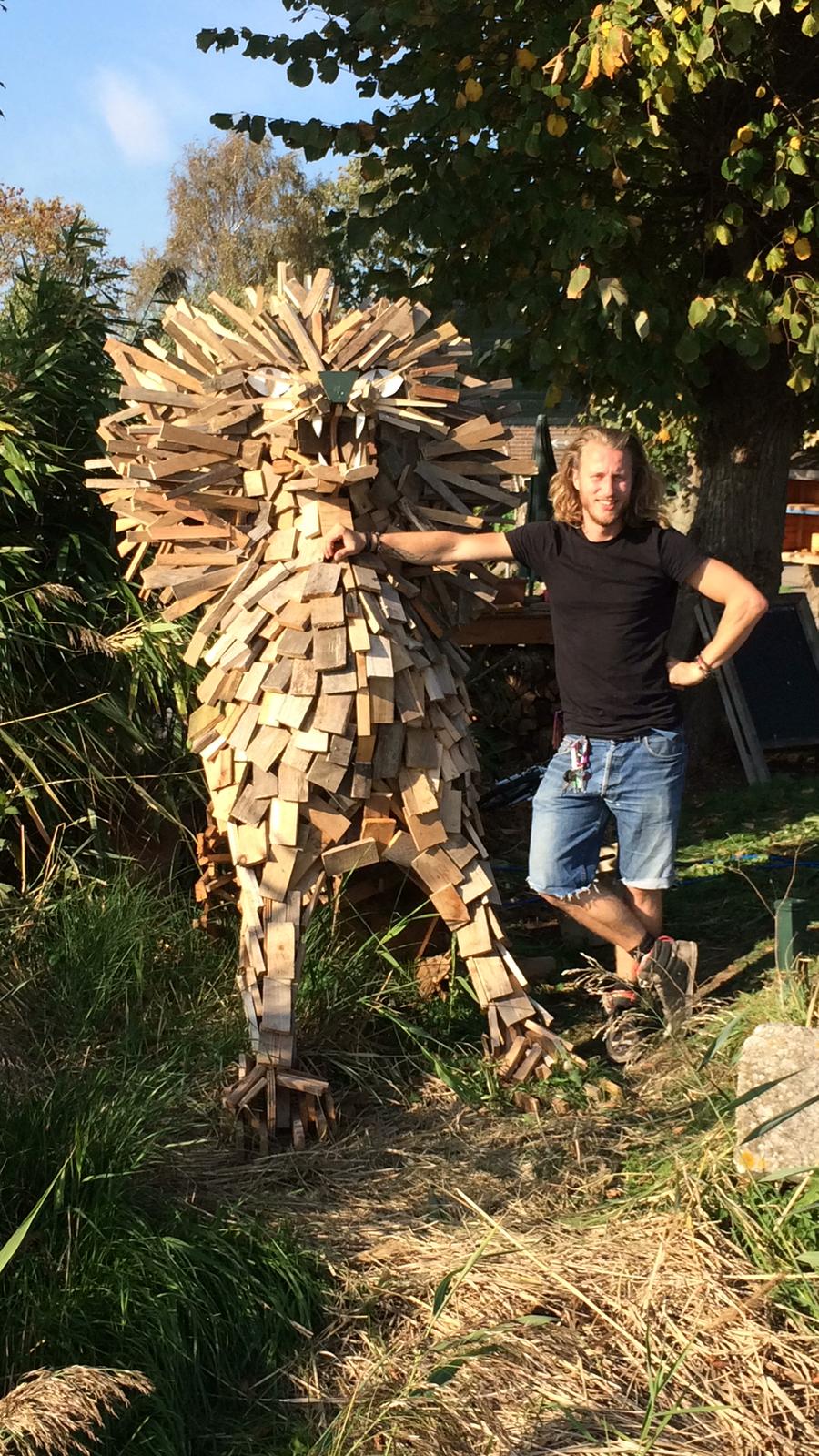 Een man staat naast een houten beeld van een leeuw, buiten in een zonnige tuin.