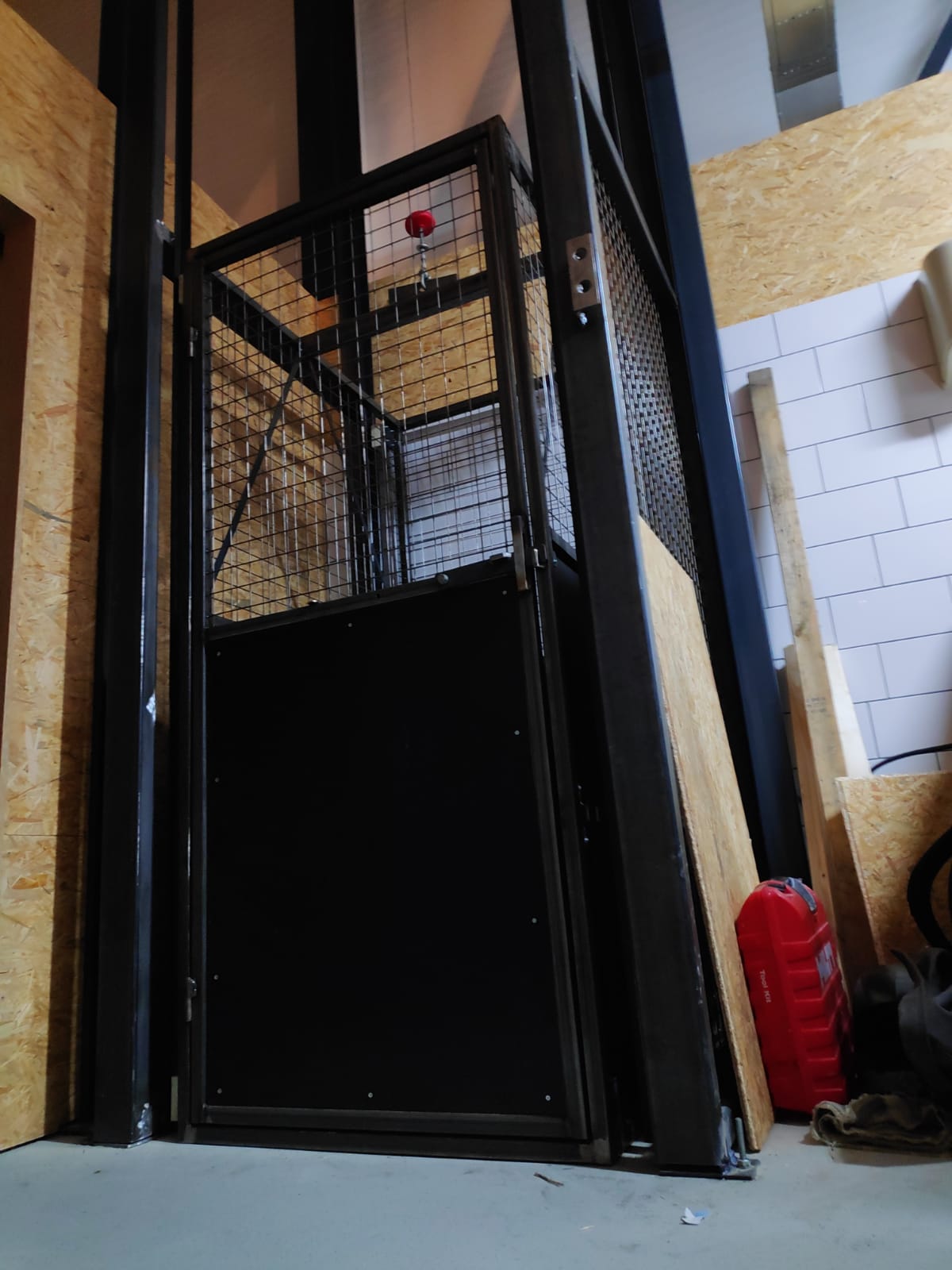 Zwarte lift met een metalen kooi, in een gebouw. Een rood licht is zichtbaar.