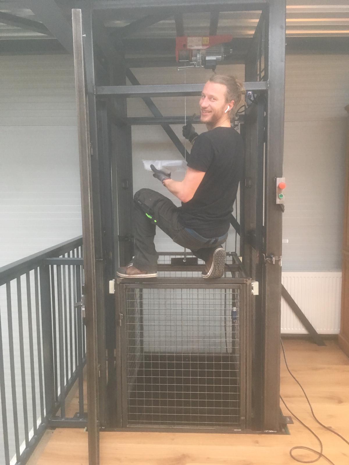 Een man zit glimlachend in een metalen goederenlift.