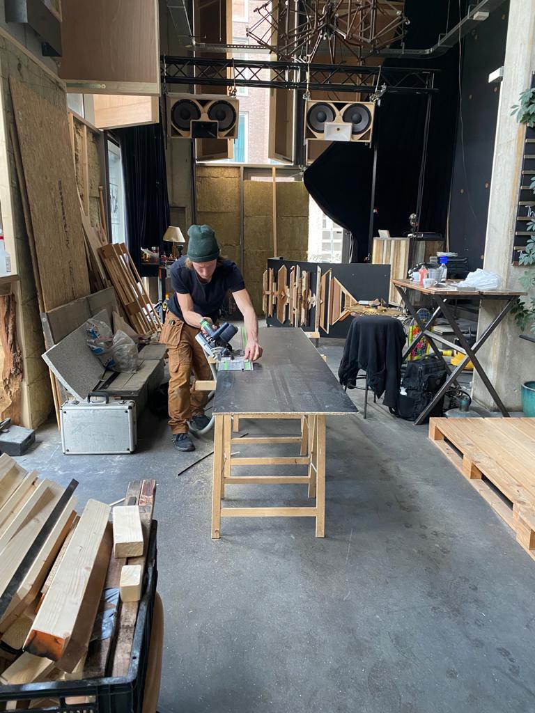 Een persoon werkt aan een tafel in een werkplaats, omringd door hout en gereedschap.