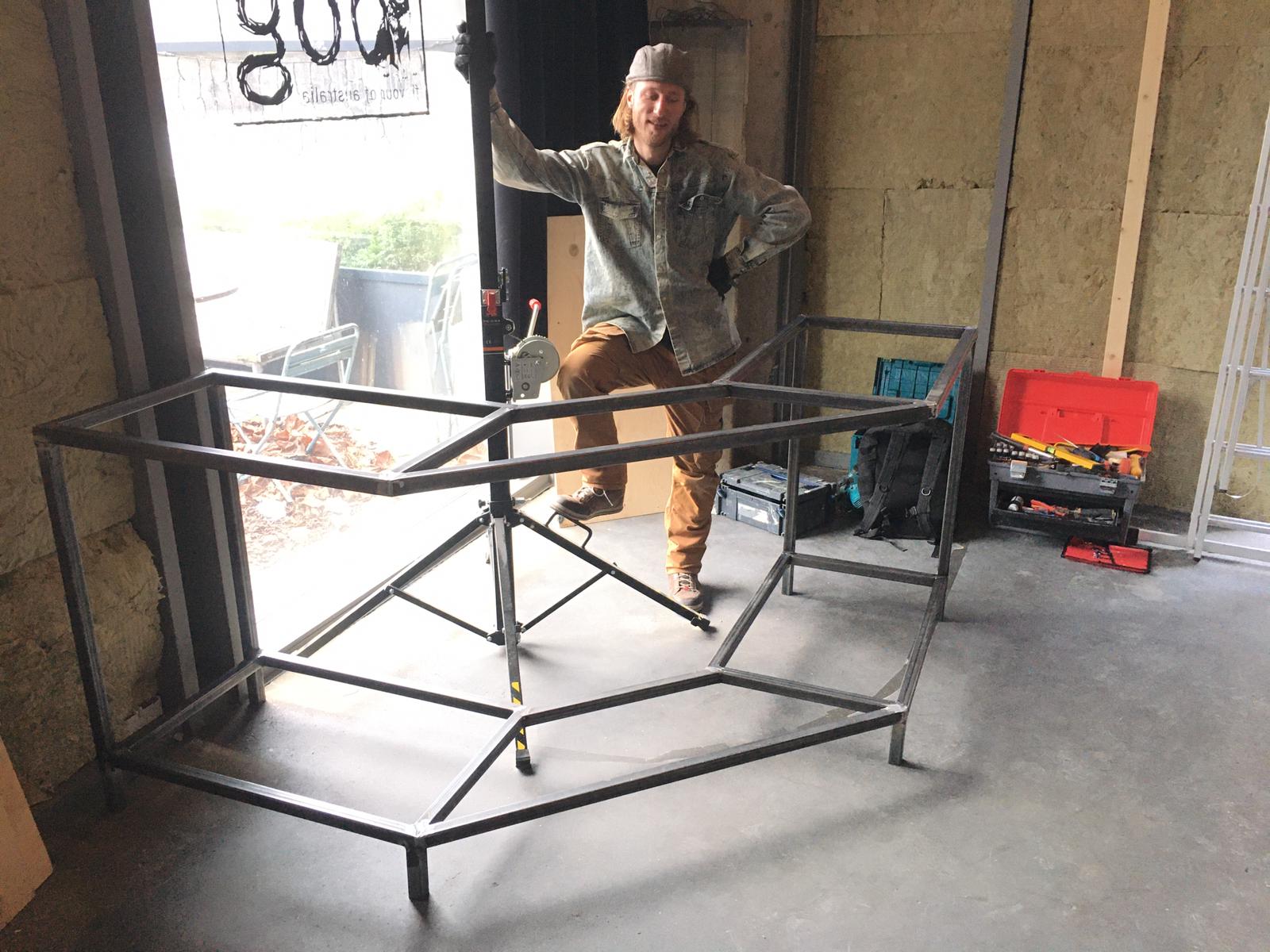 Een persoon poseert naast een groot, onafgewerkt metalen tafelonderstel met wielen in een werkplaatsomgeving.
