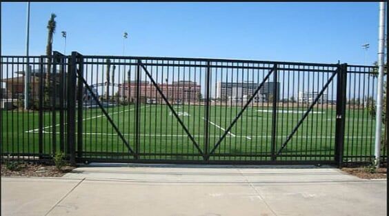 Black metal fence and gate in front of a green sports field under a clear blue sky