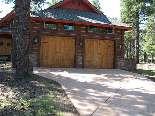 Wooden two-car garage with red trim and concrete driveway in a wooded setting