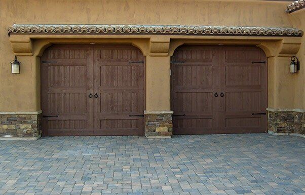 Two brown wooden garage doors on a beige stucco house with stone bases and a paved driveway