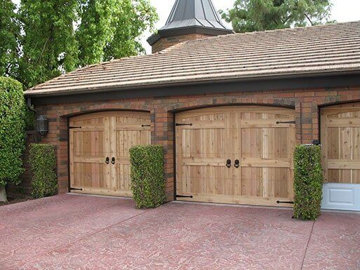 Three light wood garage doors on a brick building with a red paver driveway and trimmed shrubs.