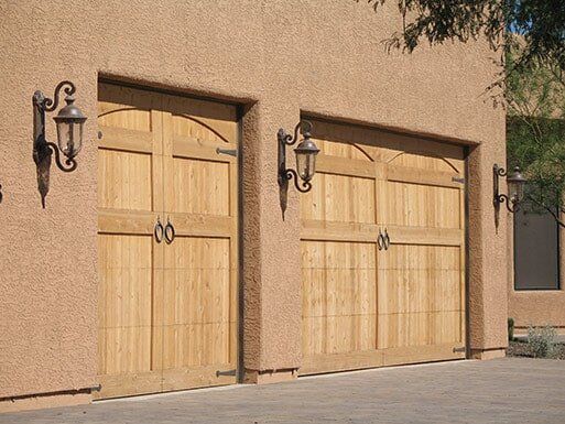 Two light wooden garage doors set in a stucco wall with black lanterns on either side