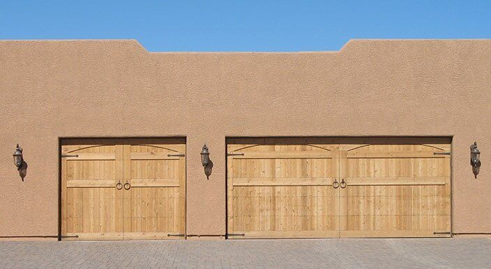 Two wooden garage doors set in a tan stucco wall under a clear blue sky.