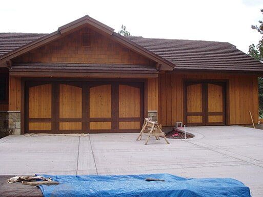 Wooden house with a tiled roof and garage doors, with construction materials on a concrete driveway.