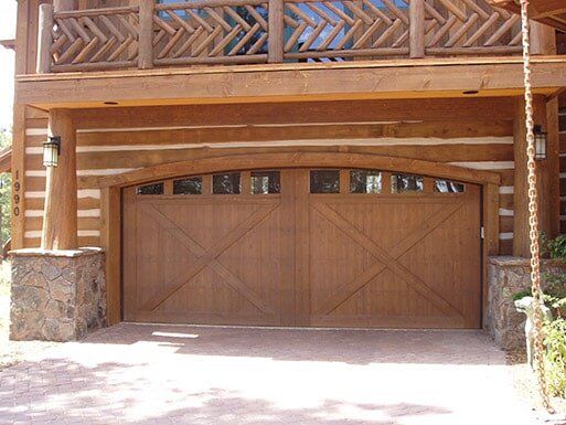 Wooden double garage door beneath a rustic balcony, with stone pillars and bright sunlight on the driveway.