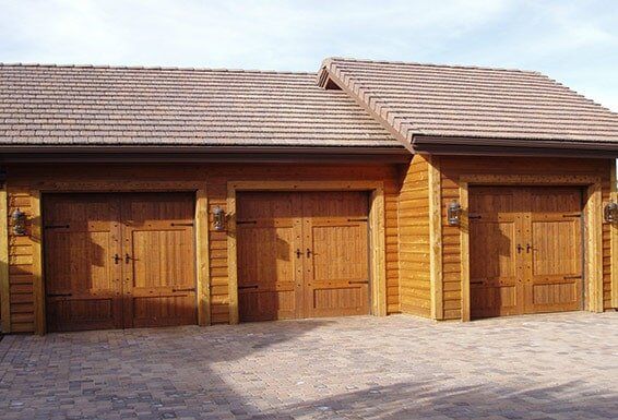 Three wooden garage doors on a tan brick building with a tiled roof and paved driveway