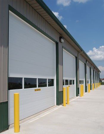 Commercial Overhead Door System — Guaranteed Door Service, Inc. Industrial warehouse units with white roll-up doors, yellow bollards, and a concrete lot under a blue sky