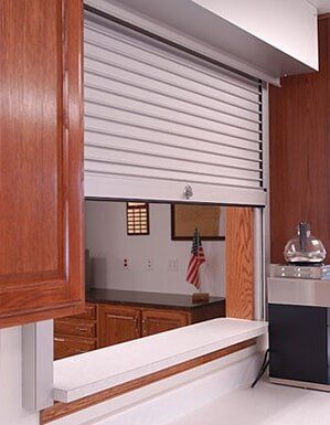 Closed white serving window with a small opening into a wood-paneled room and flag in the background