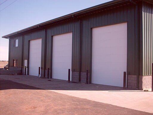 Industrial building with four large white garage doors and dark metal siding along a paved lot