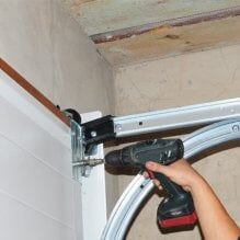 Hand holding a cordless drill fastening a garage door track bracket near the ceiling