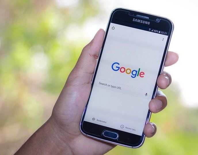A person is holding a cell phone with the google search engine on the screen.