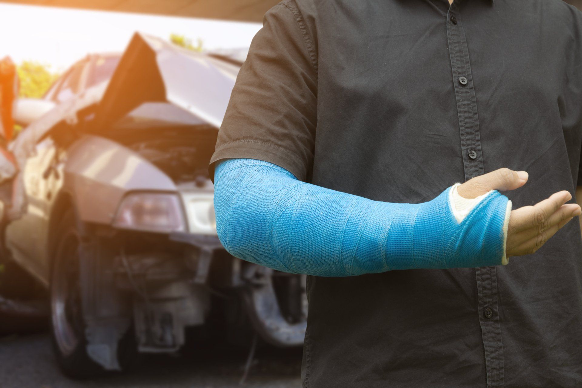 Person with blue arm cast stands in front of a damaged car.