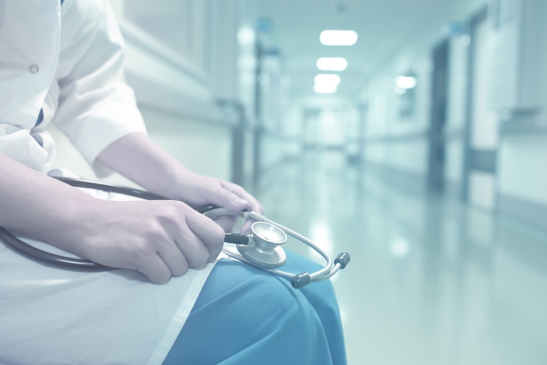 Doctor in white coat and blue scrubs holds a stethoscope, seated in a hospital hallway.
