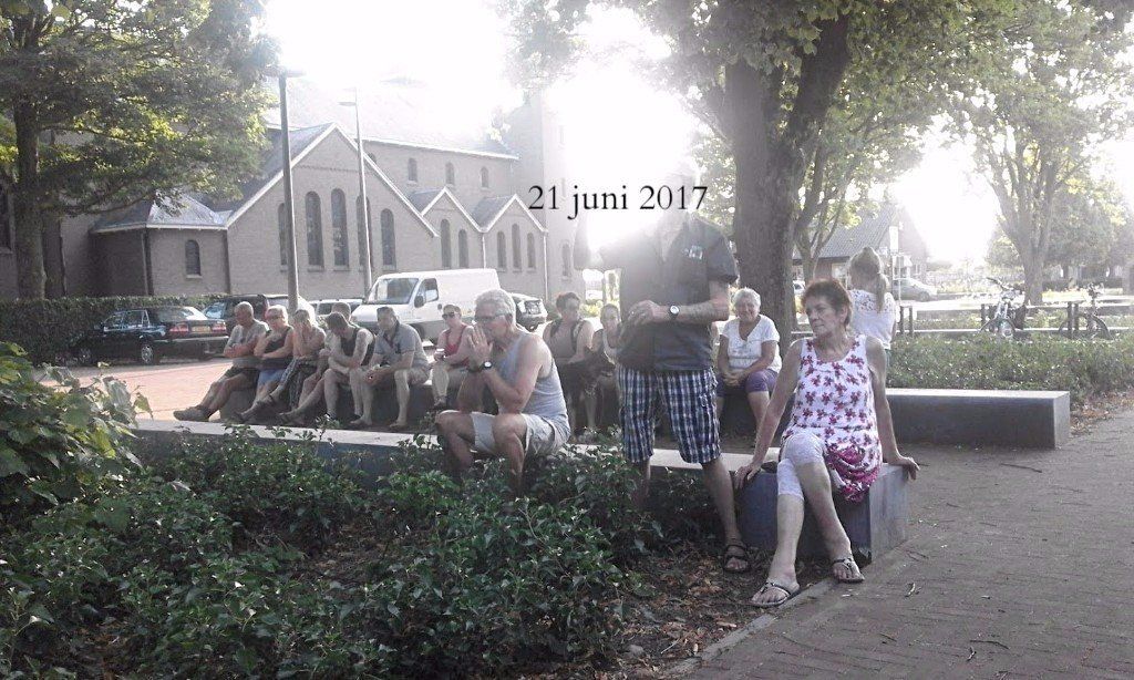 People sit on a bench in a park. A church and parked vehicles are visible in the background.