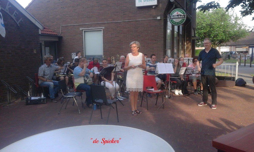 Band performs outdoors, a woman speaks. Brick building backdrop, audience seated.