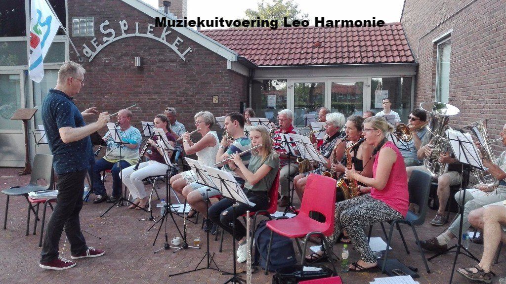 Outdoor band performance; conductor facing musicians, sheet music stands, brick building.