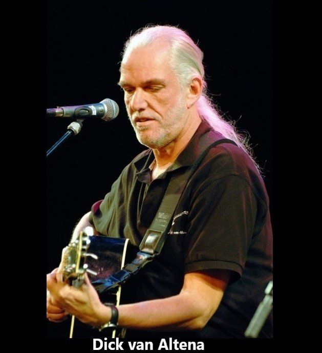 Man with white hair and beard plays acoustic guitar on stage, microphone nearby.