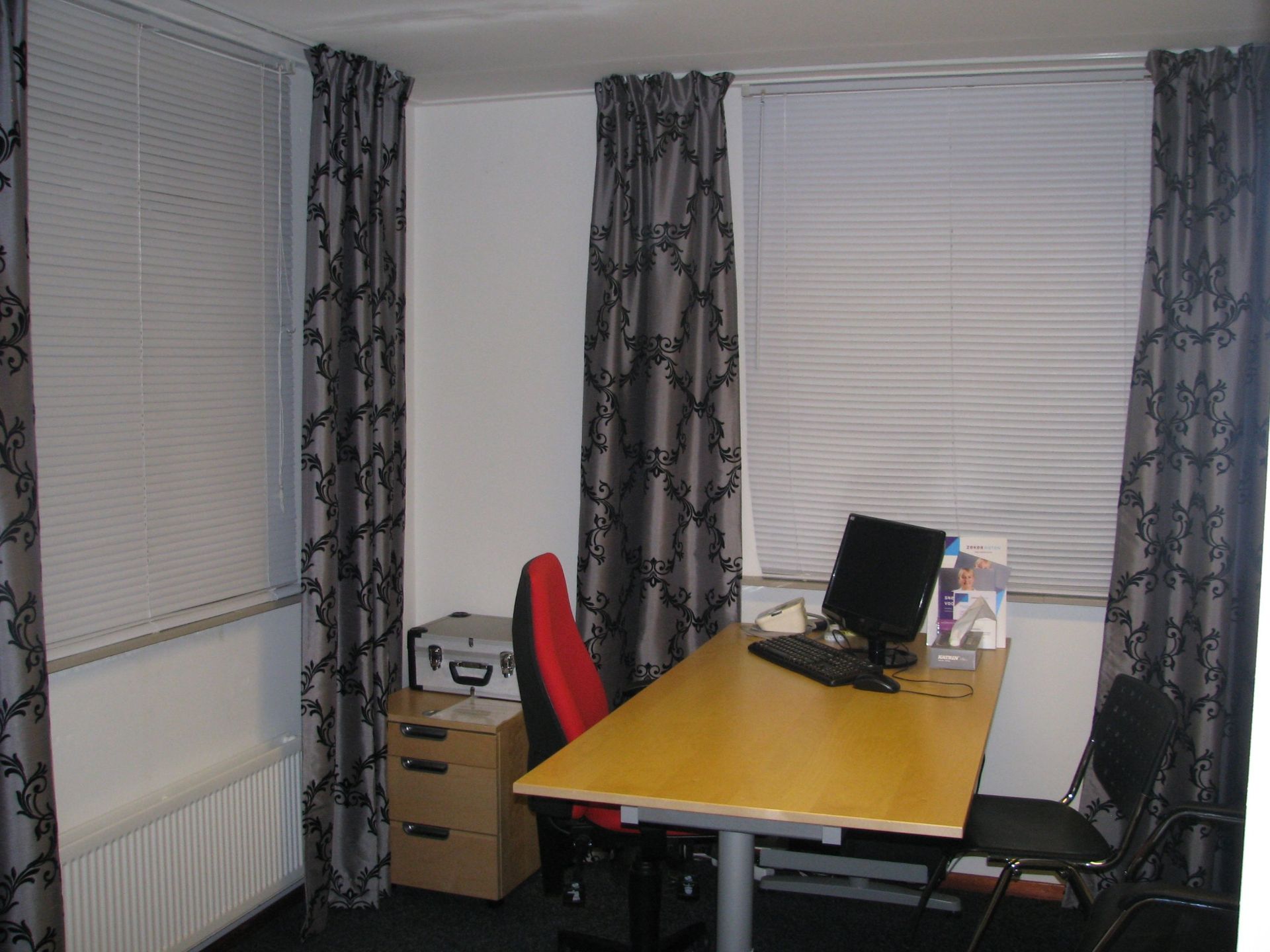 Spreekkamer; Office with desk, computer, file cabinet, and two chairs. Windows with blinds and patterned curtains.