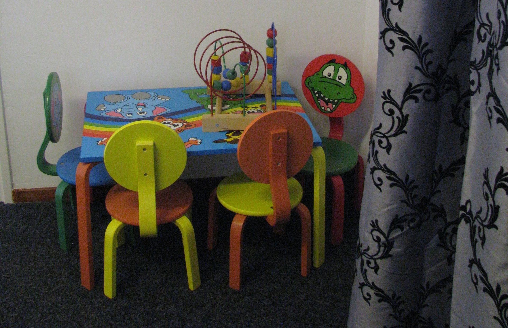 Kinderhoek dokterspost; A small, colorful children's table and chairs with toys on it, next to a patterned curtain.