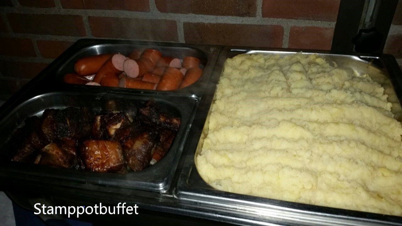 Buffet with sausage, pork, and mashed potatoes in metal trays against brick wall.