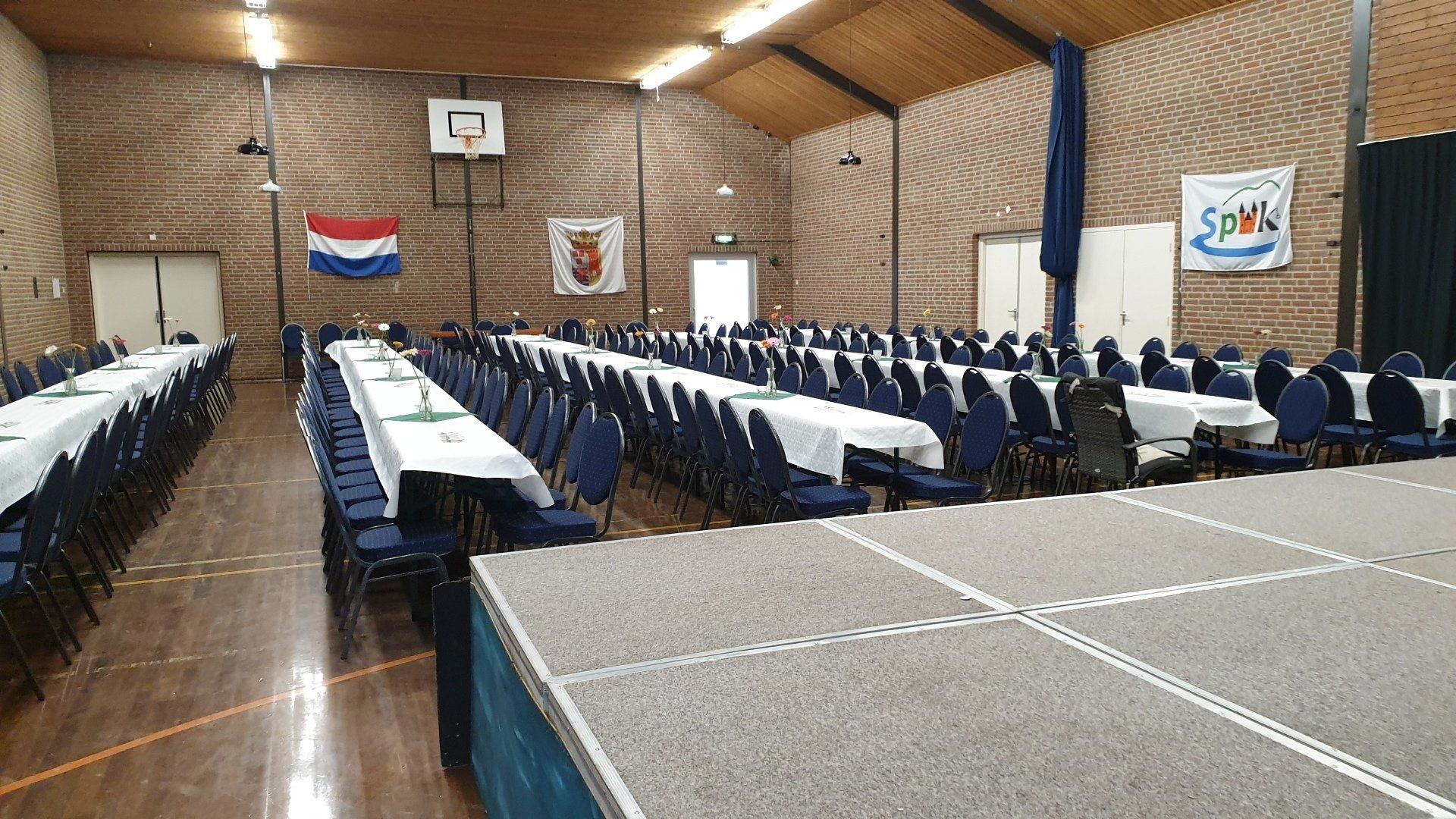 Spiek Steenghoed; Hall set up with rows of tables covered in white tablecloths and blue chairs. A stage is in the foreground.