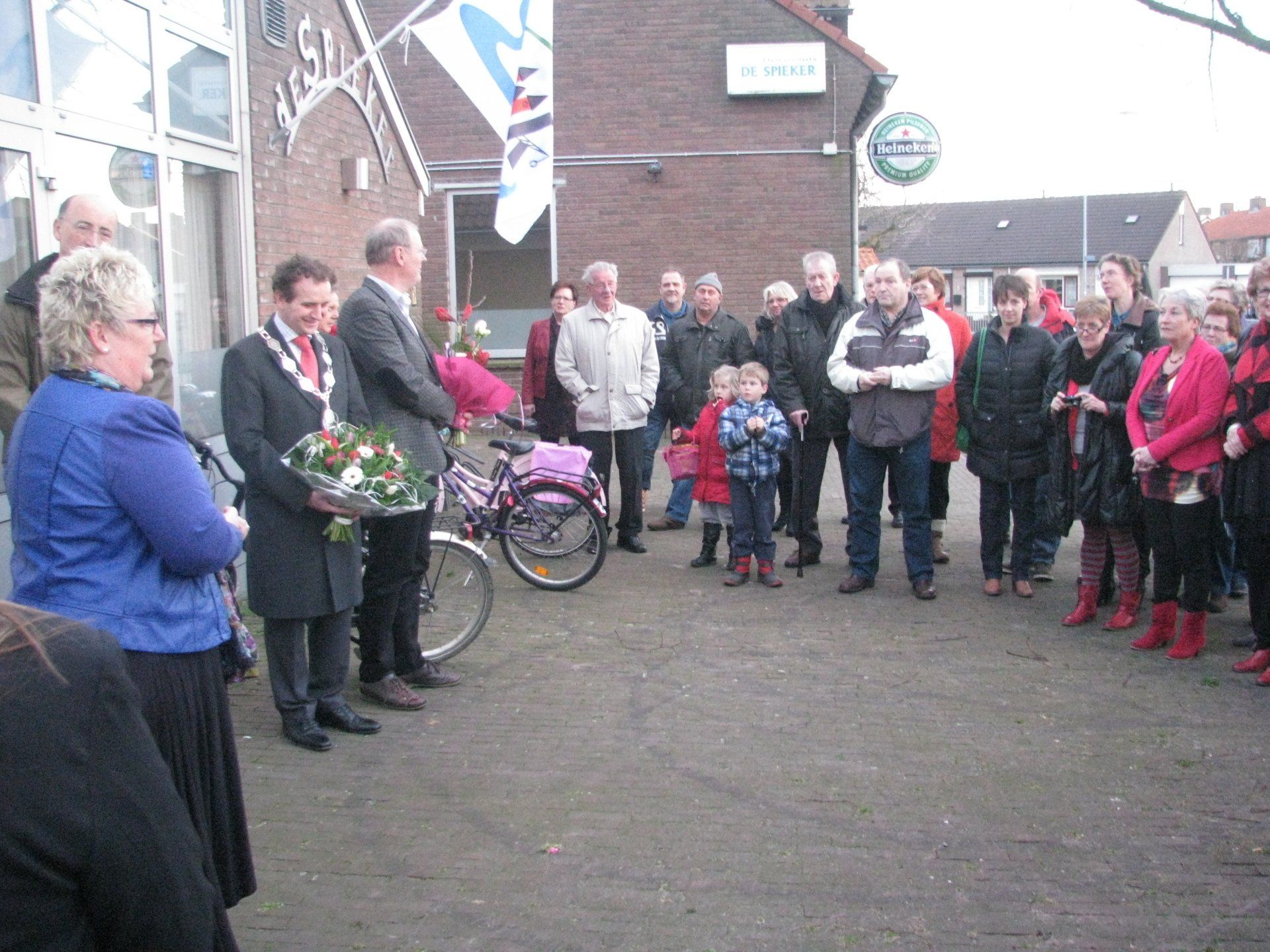 Opening dokterspost; People gathered outdoors; a man and a woman hold flowers by a bicycle, others stand around.